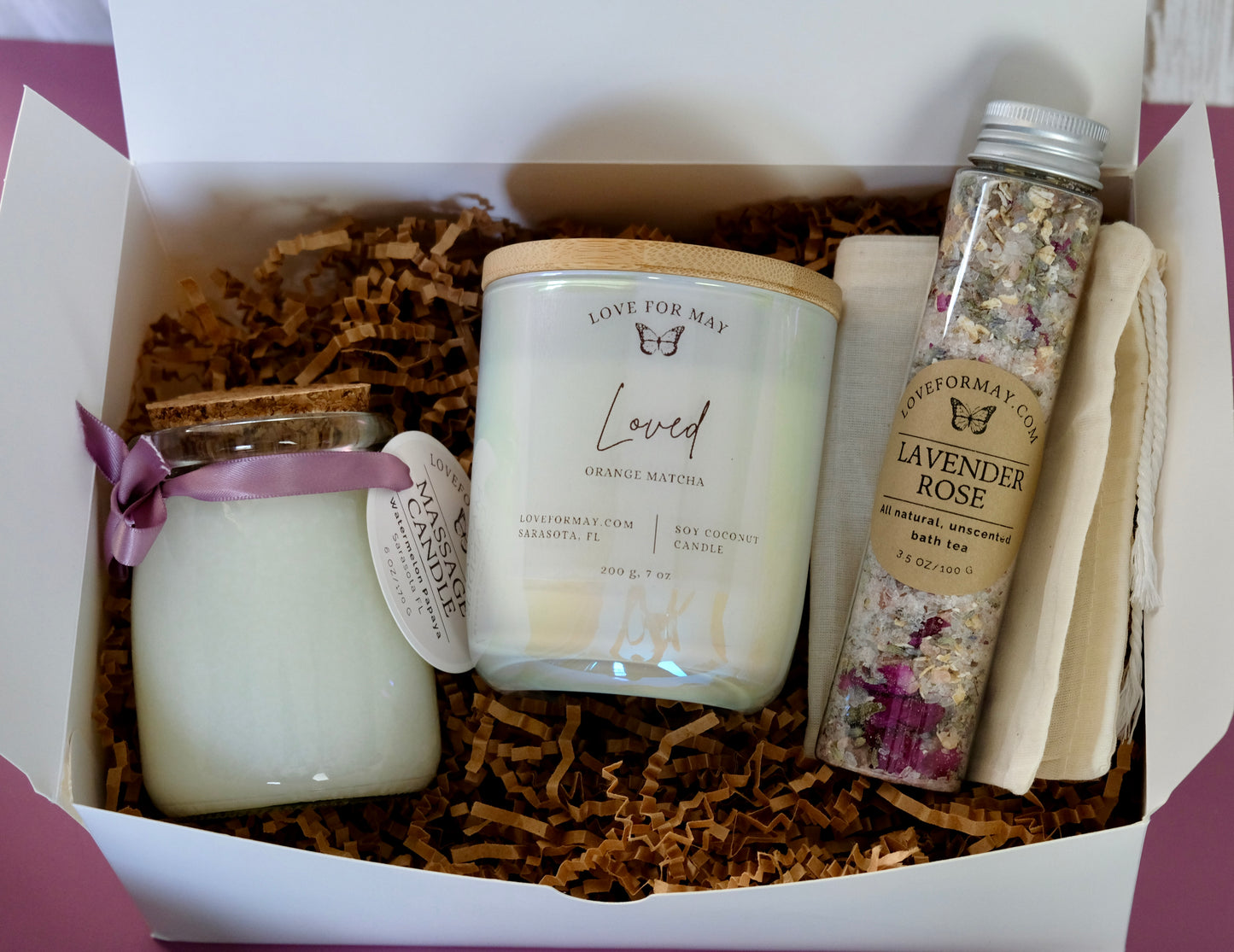 A gift box with a tube of rose lavender bath tea and bath salts, an iridescent manifestation candle, and a body oil candle. The candle says Loved.