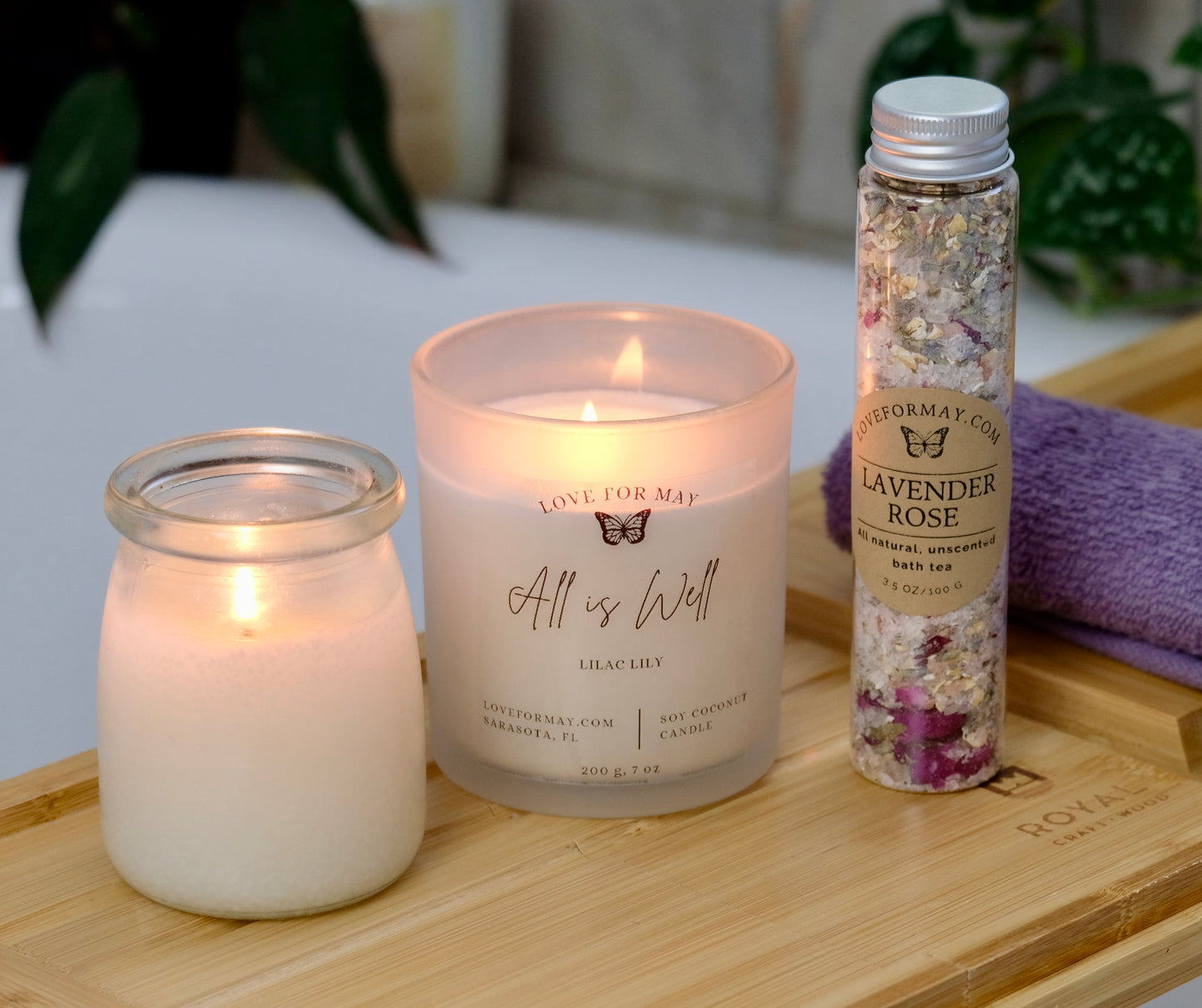 A tube of rose lavender bath tea and bath salts, an iridescent manifestation candle, and a body oil candle. The candles are lit and the products are displayed on a bath caddy.