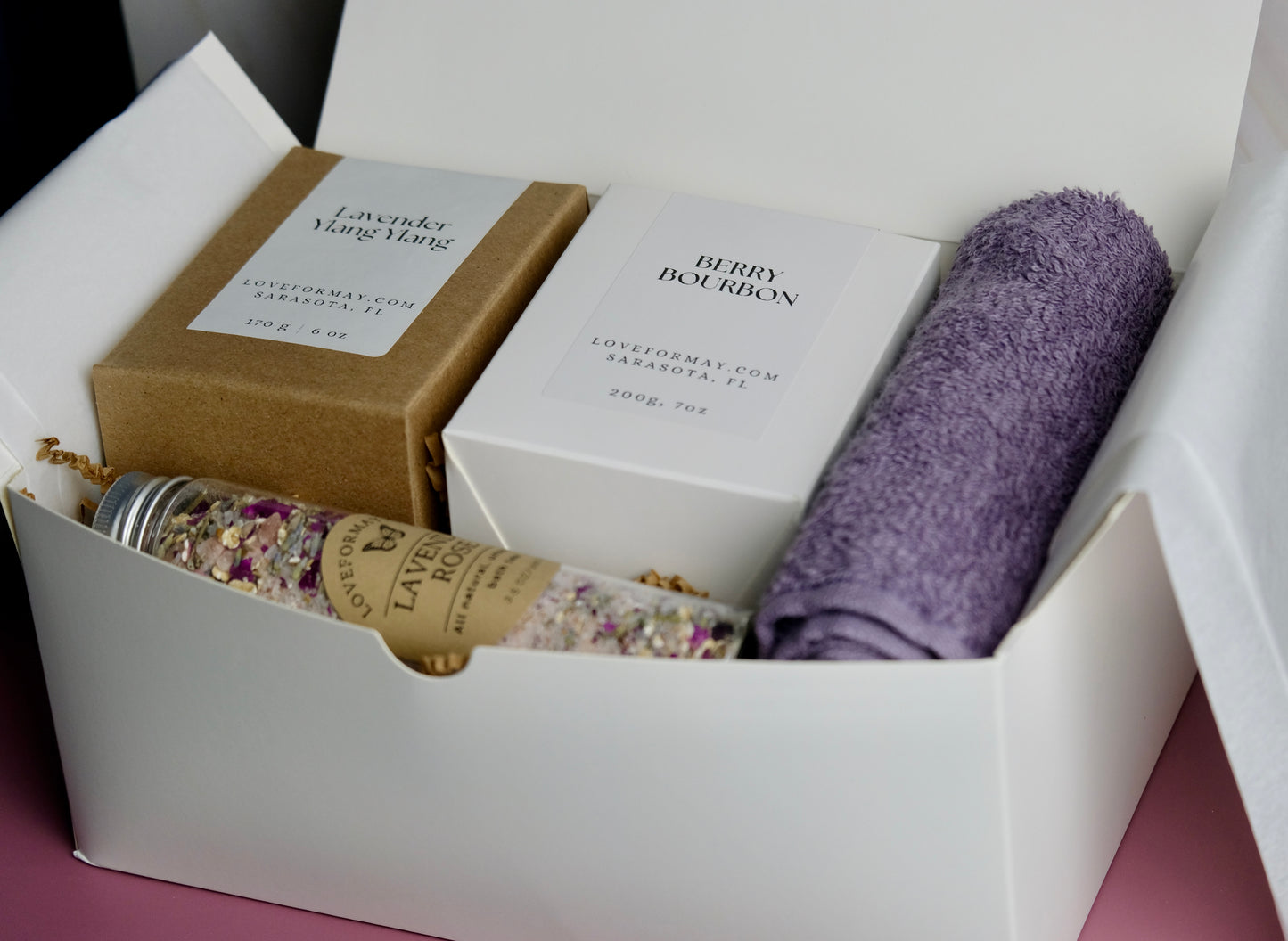 Gift box with boxed up candles, a tube of bath tea, and a purple wash cloth.