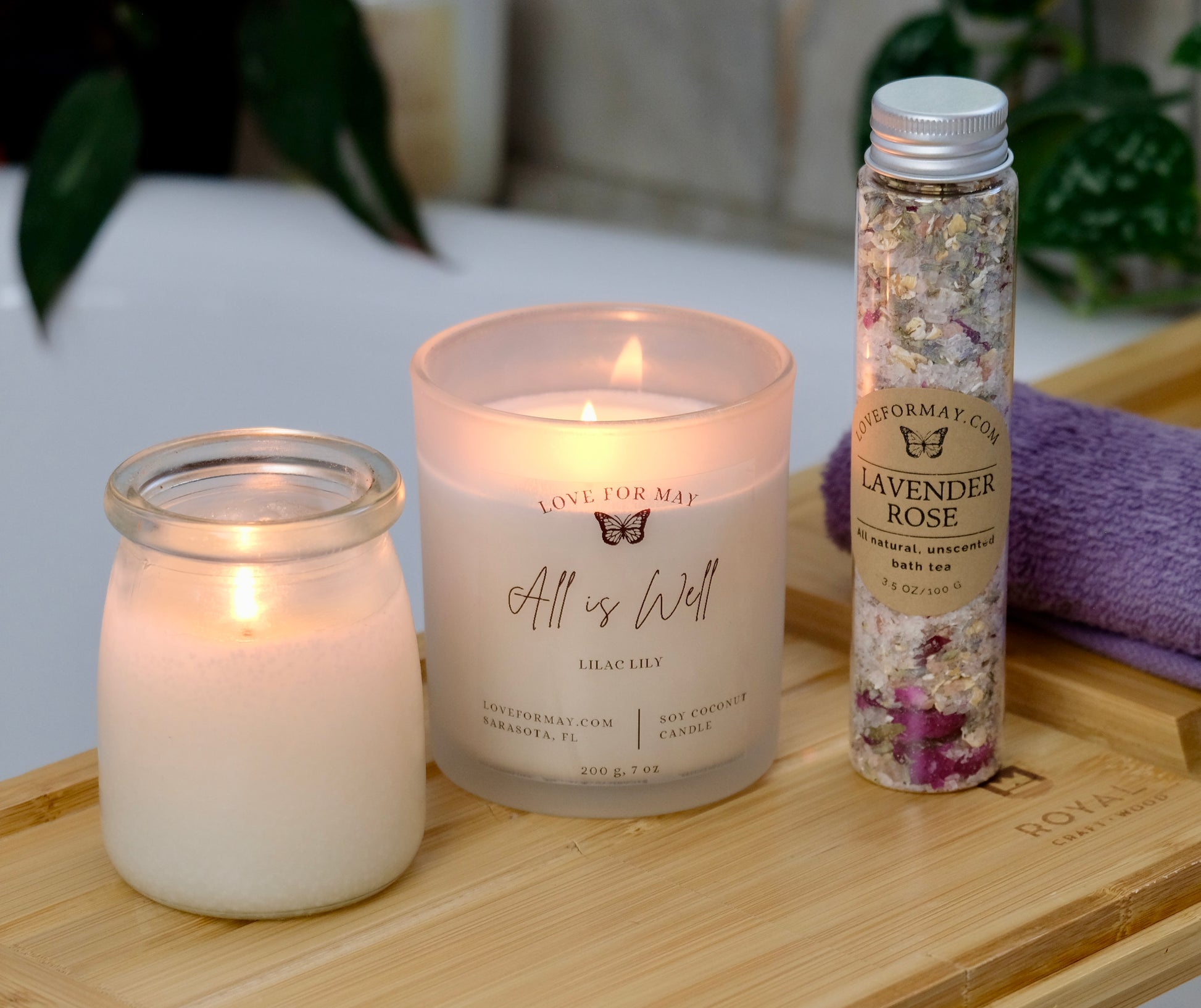 A tube of rose lavender bath tea and bath salts, an iridescent manifestation candle, and a body oil candle. The candles are lit and the products are displayed on a bath caddy.