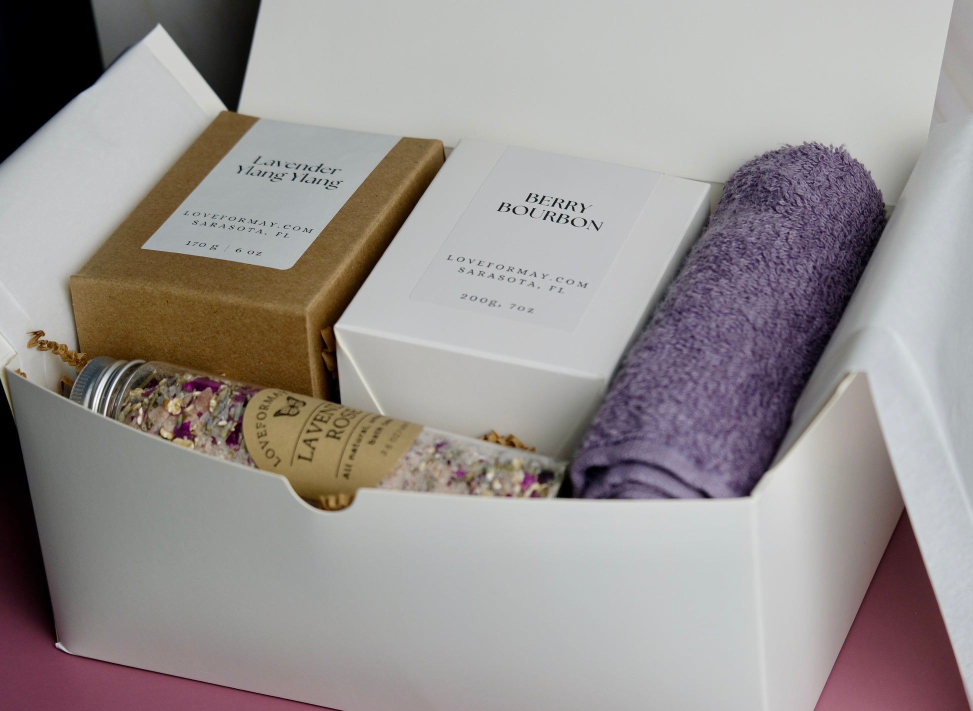 Gift box with boxed up candles, a tube of bath tea, and a purple wash cloth.