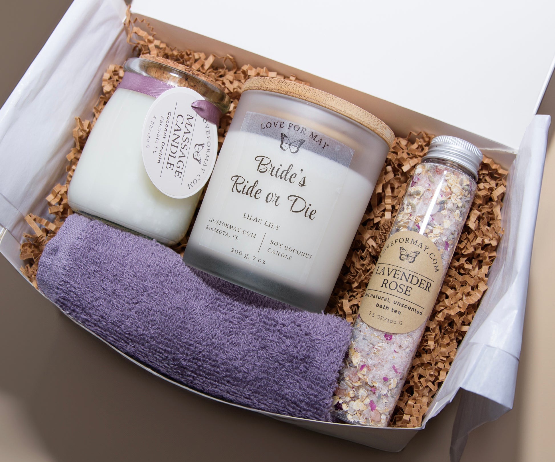 A gift box with a massage candle, a personalized handmade candle that says Bride's Ride or Die, bath tea, and a purple washcloth.