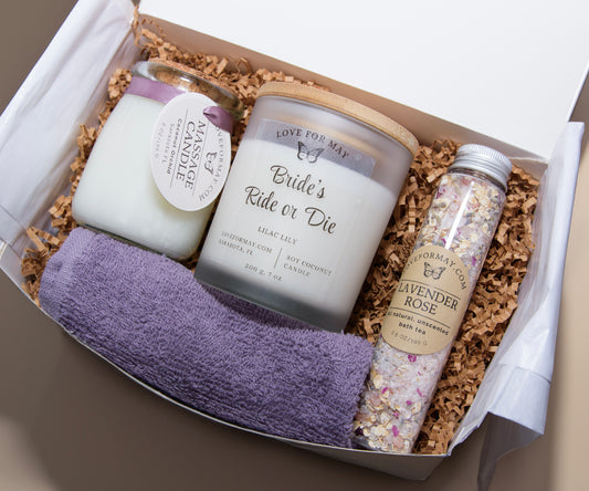 A gift box with a massage candle, a personalized handmade candle that says Bride's Ride or Die, bath tea, and a purple washcloth.