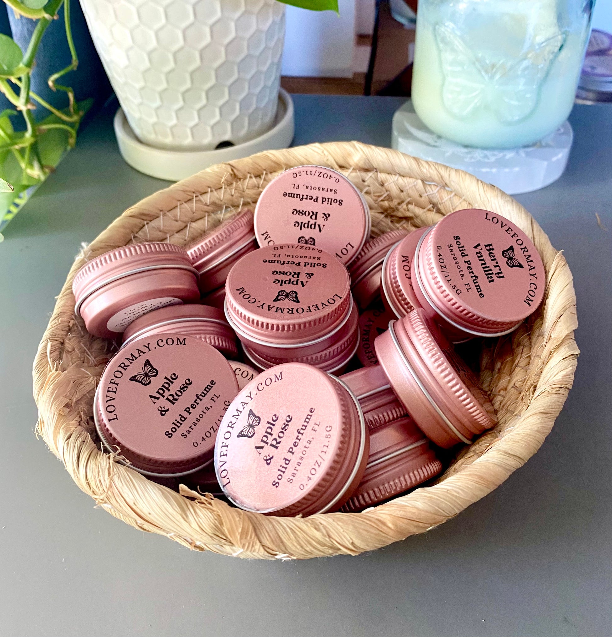 Basket of pink solid perfume tins with butterfly branding on a gray surface.