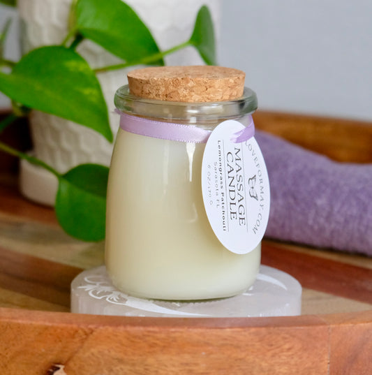 Massage candle in a glass jar with cork lid on a wooden surface, surrounded by green leaves and a purple towel.