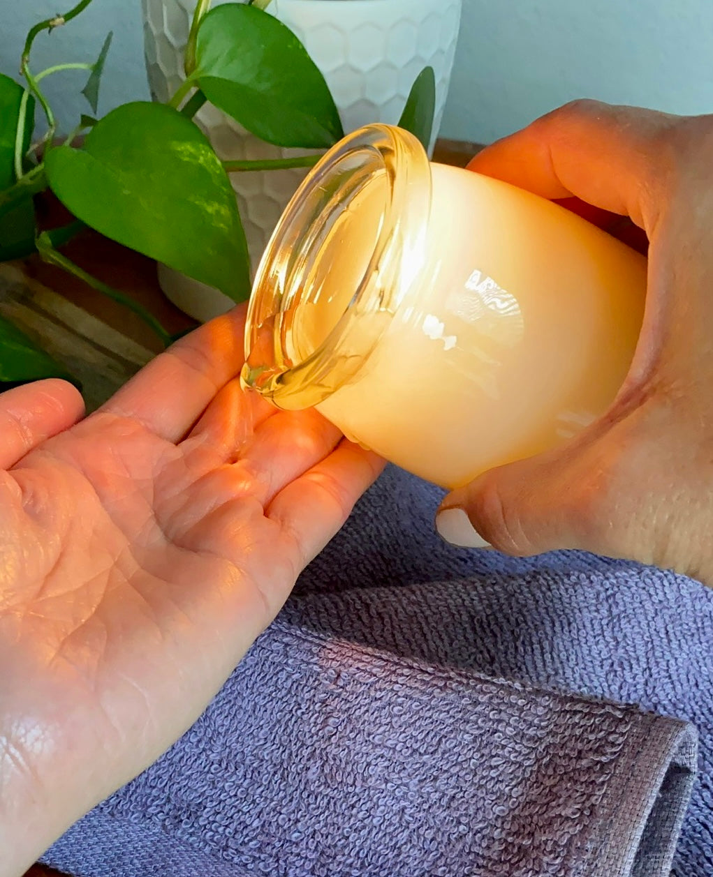 Hand pouring oil from a massage candle into hands, on a purple towel with green leaves in the background.