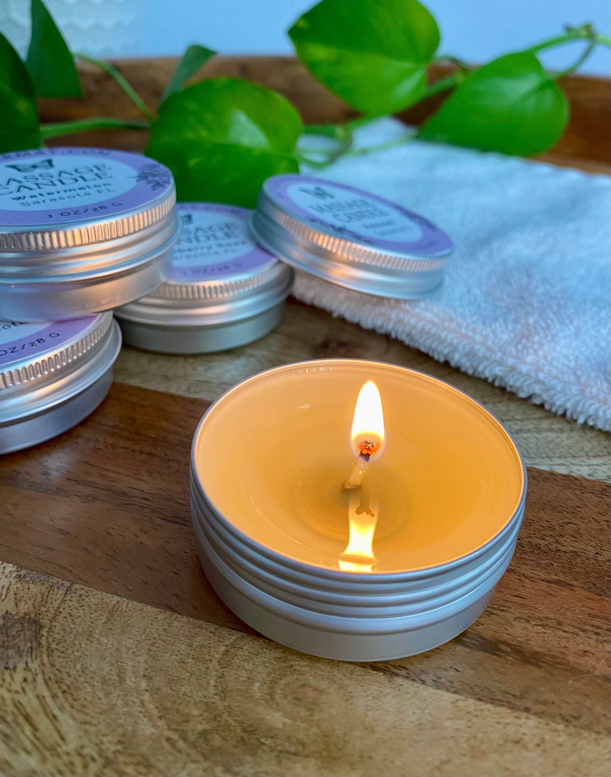 Stack of small massage candles with a lit candle on a wooden surface, surrounded by green leaves and a white towel.